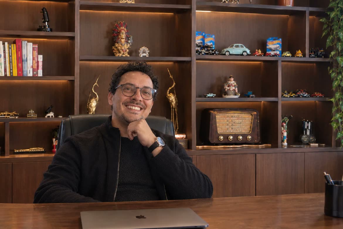Wagner Borges de Lima sorrindo, sentado à mesa do escritório, com uma estante de madeira iluminada ao fundo.